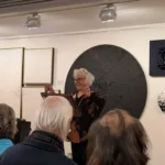 GACL-President-Cathy-Alexopoulos-addresses-audiences-at-the-Steps-Gallery-standing-in-front-of-works-by-Victoria